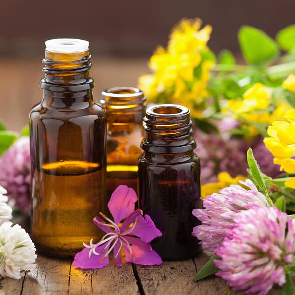 Three glass bottles of herbal extracts and essential oils surrounded by vibrant flowers, placed on a rustic wooden table.